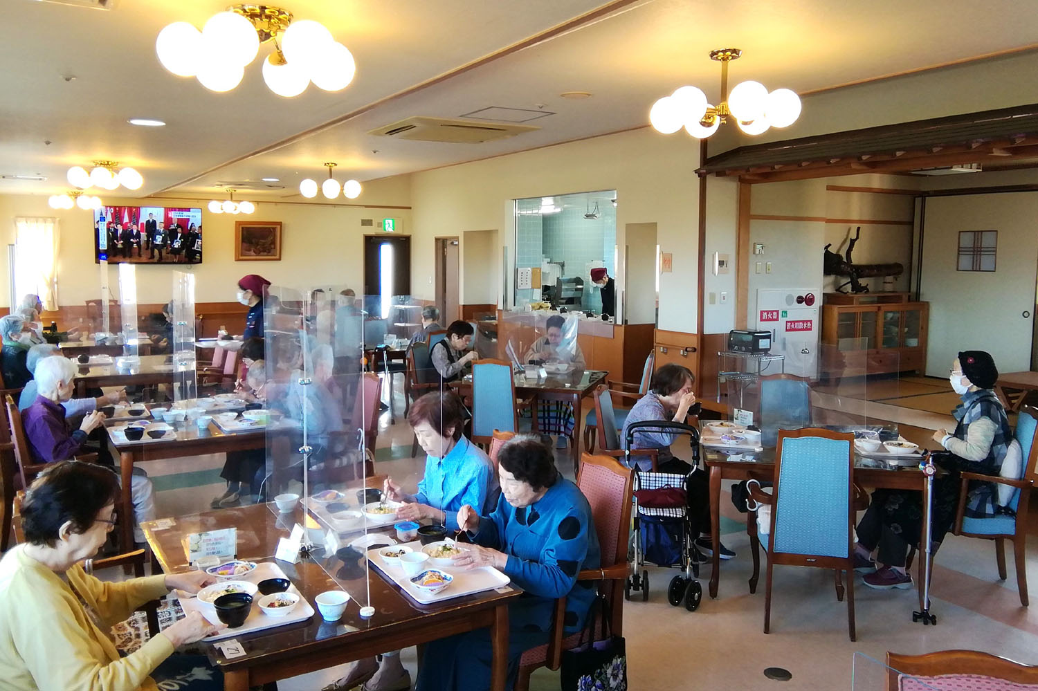 介護施設の食堂。高齢者が食事をしており、スタッフが配膳対応。テーブル間にアクリル板設置。